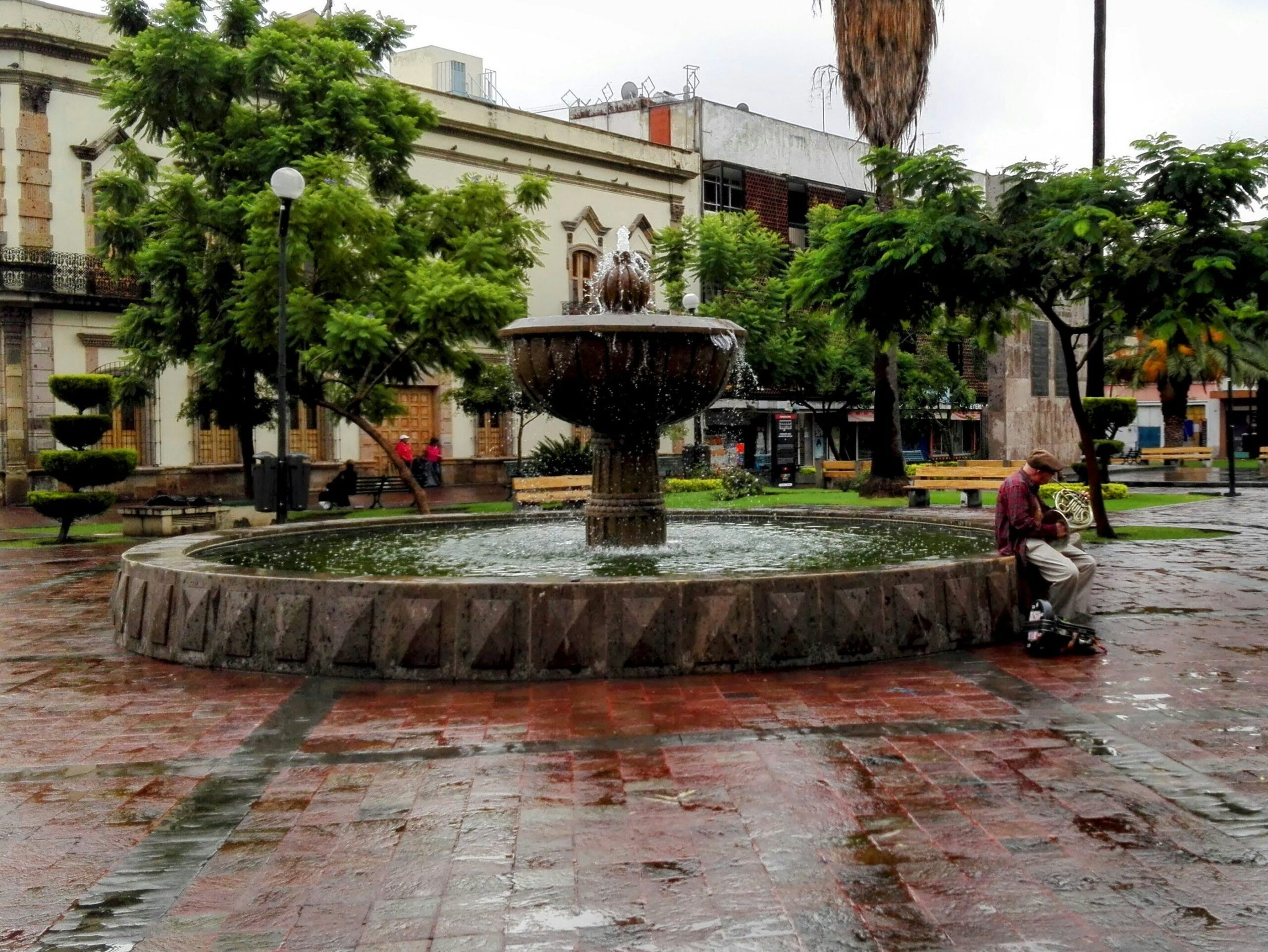 Fuente central en plaza con agua brotando y un músico sentado tocando un corno junto al borde bajo la lluvia
